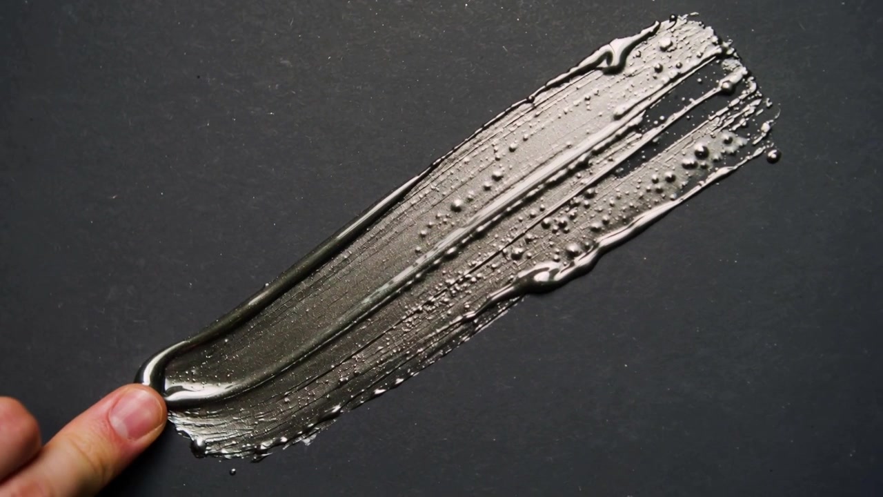 Yema del dedo tocando, presionando y arrastrando gel metálico sobre una lámina mate oscura, revelando textura.