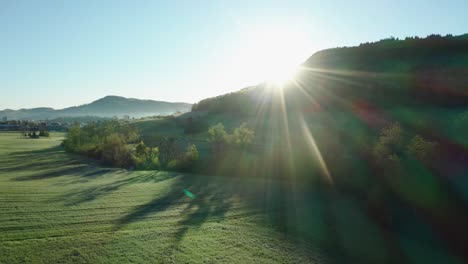 aerial-drone-view-of-morning-hour-over-peaceful-countryside-with-green-and-agricultural-fields,-sun-shining-directly-into-camera,-switzerland