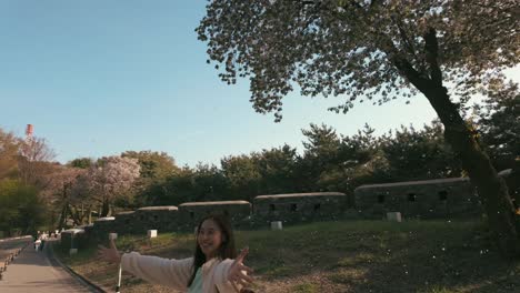 Observe-Cómo-Una-Joven-Alegre-Baila-Bajo-Una-Cascada-De-Pétalos-De-Flores-De-Cerezo,-Abrazando-La-Serena-Belleza-De-La-Primavera-En-Un-Pintoresco-Parque.