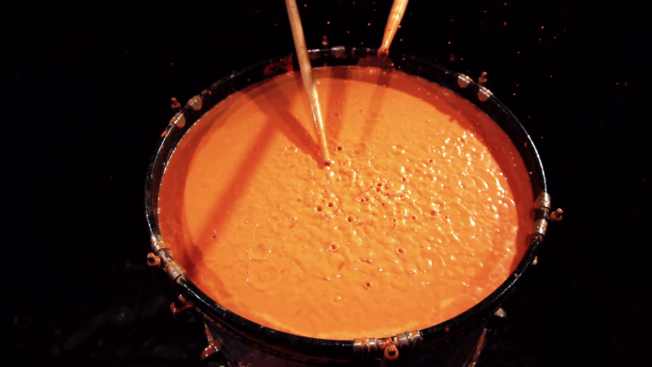 Slow motion footage of drumsticks hitting the top of a drum that is filled with orange liquid
