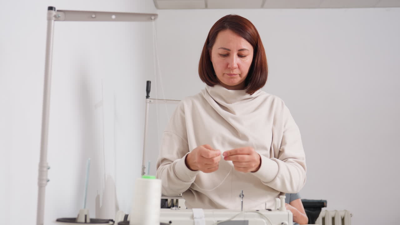 Fashion designer carefully strengthening thread on sewing machine in bright minimal workspace, focusing on precision while second person works in background, showcasing expertise, and true skill