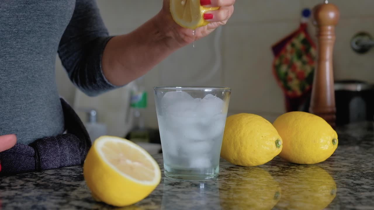 Mujer exprimiendo un limón con ambas manos sobre un vaso con hielo entre limones