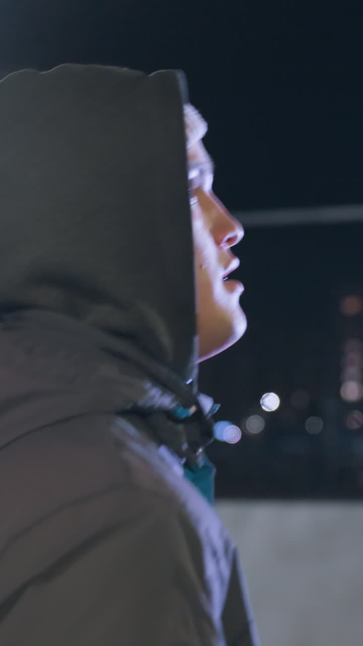 hombre con capucha hábilmente corre la pelota durante la práctica nocturna en el campo deportivo urbano, la atrapa con la mano derecha, y continúa corriendo bajo luces de calle brillantes rodeado de paisaje urbano y edificios