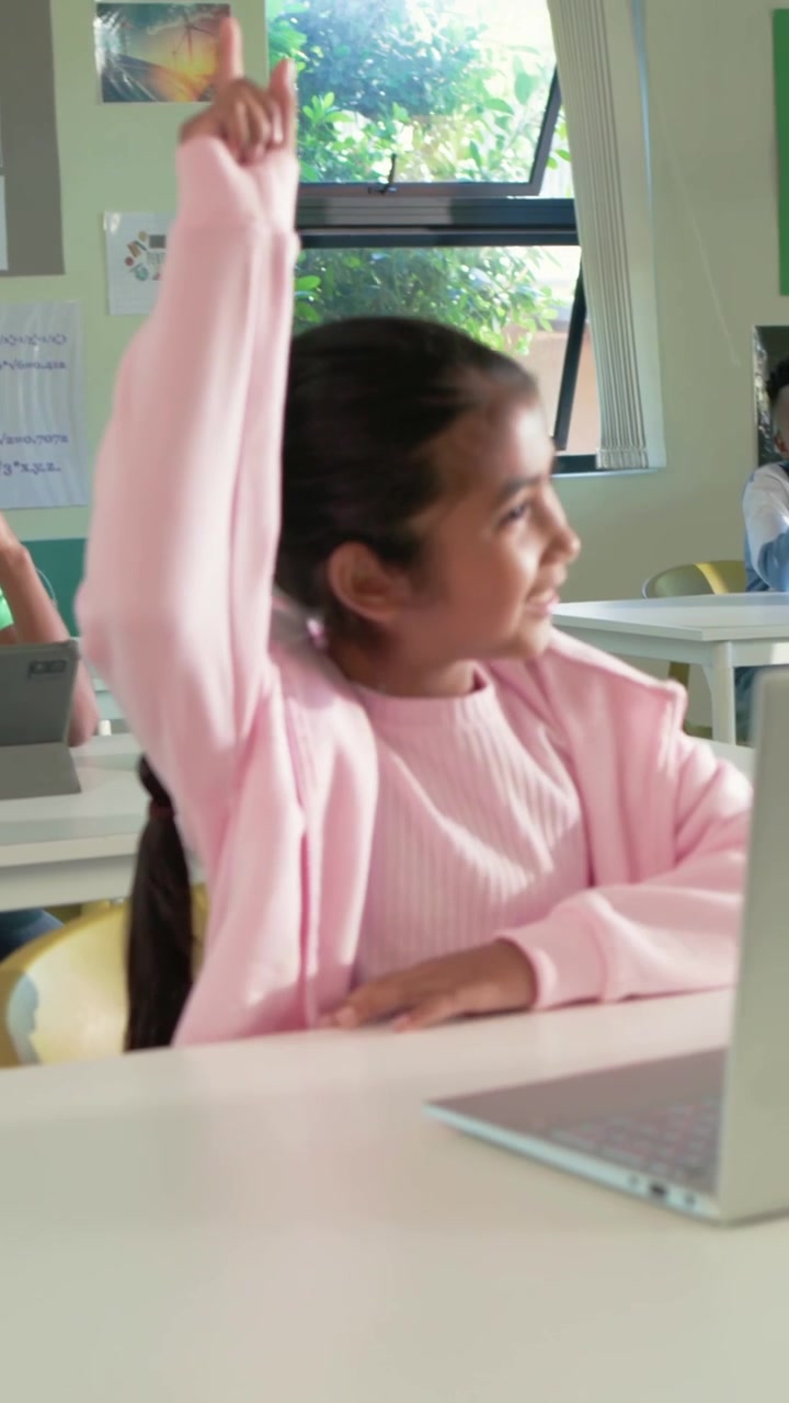 Video verticale: Ragazza con maglione rosa che alza la mano e risponde all'insegnante, seduta a una scrivania con un laptop.