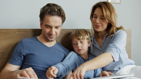 Joven-Madre-Y-Padre-Sentados-En-La-Cama-Por-La-Mañana-Con-Su-Pequeño-Hijo-Y-Leyendo-Un-Libro-Interesante