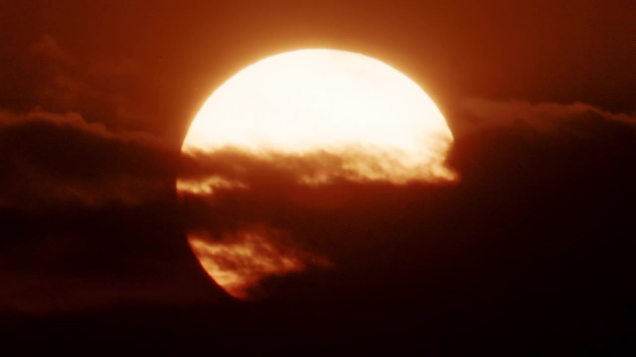Close up of a bright sun partially covered by thick moving clouds, glowing in intense golden orange sunset tones