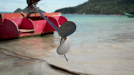 a red wooden boat with a visible propeller resting on a sunny beach, gentle waves washing ashore under a clear blue sky