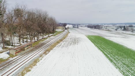 Vista-De-Ariel-De-Una-Máquina-De-Vapor-Y-Automóviles-De-Pasajeros-Resoplando-A-Lo-Largo-De-Las-Tierras-Agrícolas-Amish-Después-De-La-Primera-Nevada-De-La-Temporada