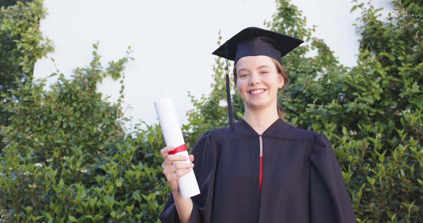 Female graduate in gown cap pulling diploma, tossing catching while celebrating at white vine wall