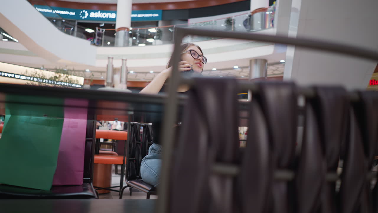 primer plano de sillas dispuestas con una dama con gafas involucrada en una llamada telefónica, disfrutando de la conversación en un centro comercial bellamente iluminado con inscripciones brillantes