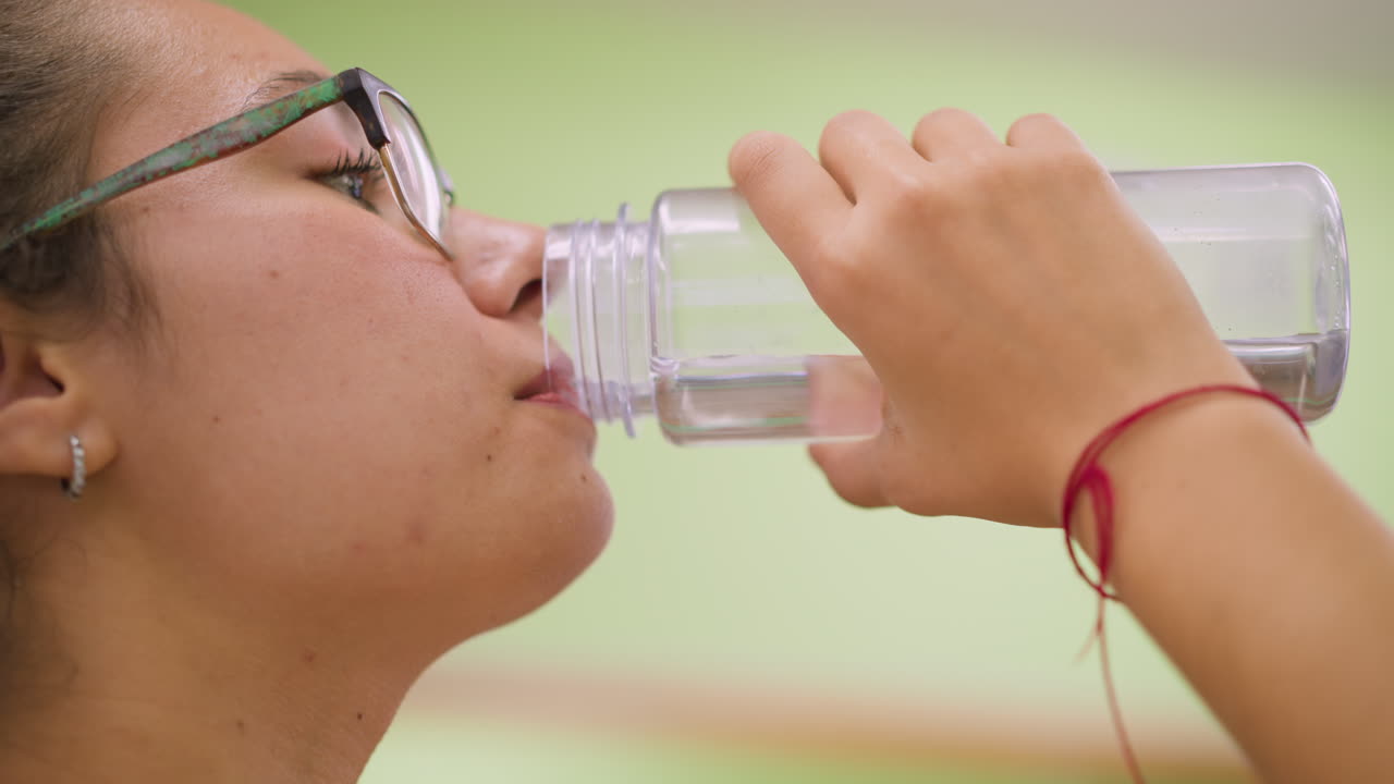 Close up view woman in white top and glasses drinking water indoors with relaxed face holding clear bottle soft natural light background showing lifestyle hydration wellness freshness beauty energy
