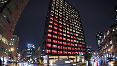 illuminated skyscraper reflecting on wet street, red and white urban lights creating vibrant nighttime cityscape with downtown architectural glow