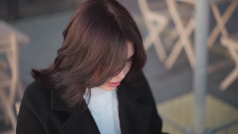 밖에서 앉아있는 어린 소녀, 그녀의 앞에 있는 무언가에 초점을 맞추고, 그녀는 그녀의 머리를 조정, 배경에 나무 의자와 기둥의 흐릿함