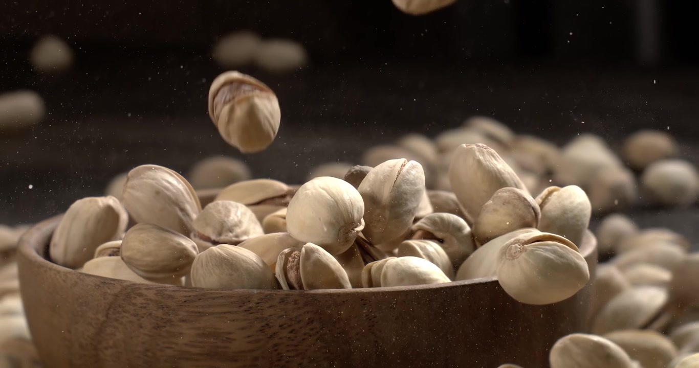 Ciotola di legno piena di pistacchi su un tavolo di legno scuro, gustosi pistacchi salati sparsi intorno, concetto di spuntino sano e cibo naturale. Ripreso con telecamera super slow motion a 1600 fps.