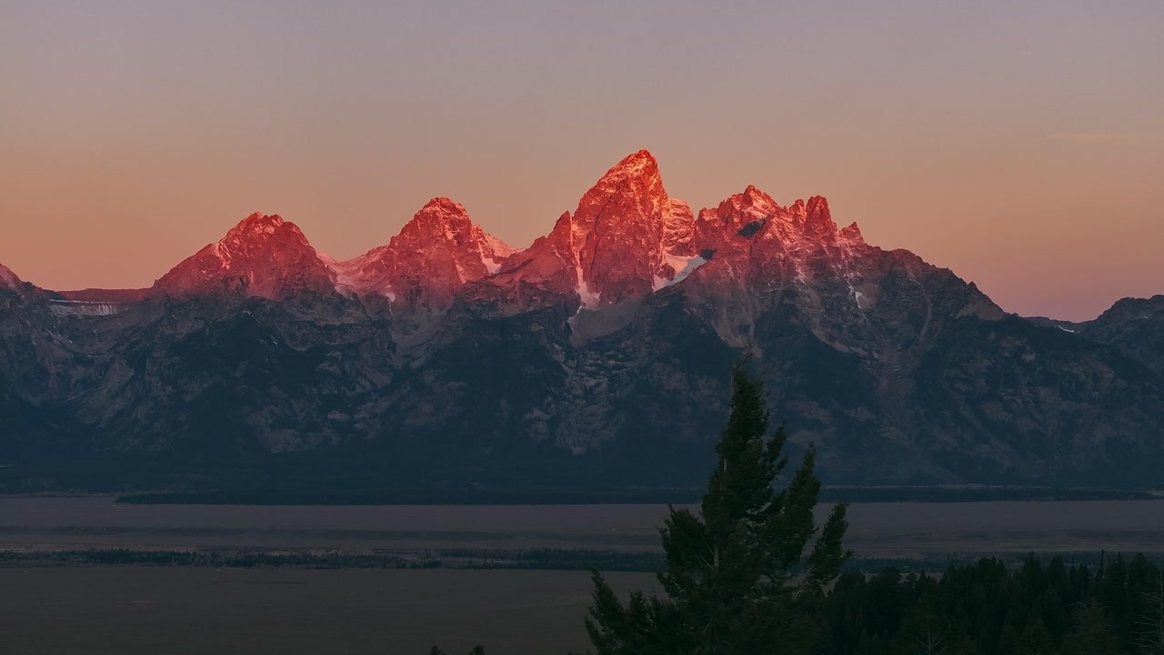 Sunrise sunset golden alpenglow on snowy Grand Teton mountain peaks above valley, aerial telephoto establishing backdrop