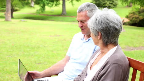 retired couple sitting on a park bench using a laptop