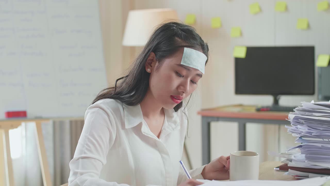 병든 아시아 여성이 사무실에서 문서와 함께 열심히 일하면서 물을 마시고 있습니다.