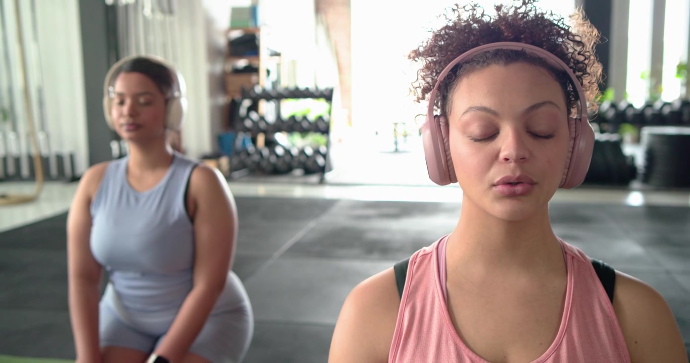 Camera panning revealing diverse female friends with headsets breathing mindfully in squat at gym