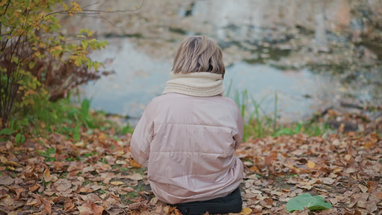 back view of solitary lady wrapped in soft scarf sitting calmly on leafy forest ground facing serene lake surface, surrounded by autumn colors and tranquil atmosphere in peaceful outdoor setting