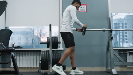 young student carrying metal weight plate across gym floor to fix onto barbell rack by window showing residential building outside, showcasing hands, gym equipment, rubber mat and focused training