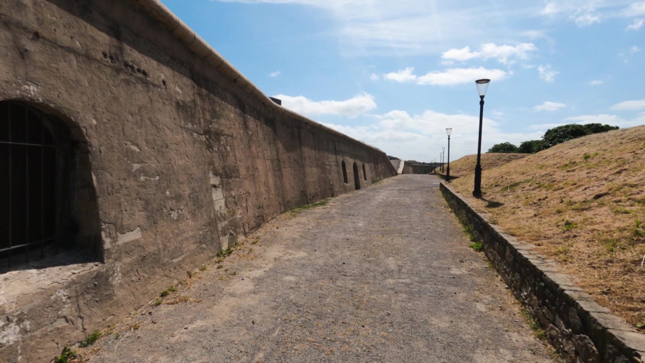 Un sentiero di ciottoli corre lungo le massicce mura della fortezza di artiglieria a Cerro Santa Catalina, Gijon. Cielo sereno, collina erbosa.