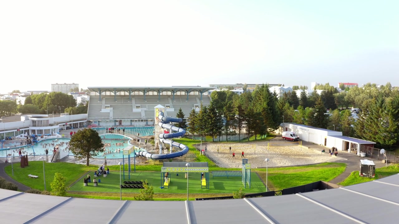 A rising aerial shot reveals a large blue water slide and swimming pool complex behind a modern white building in Reykjavik, Iceland on a bright day.