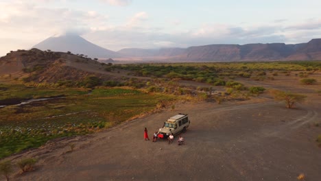 an amazing drone shot of a safari jeep parked on a hilltop with people sitting outside and enjoying the sunrise view of lake natron and ol doinyo lengai volcano in the background in tanzania in africa