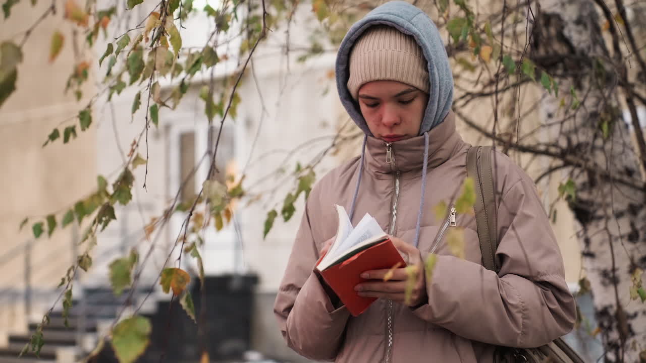 Light skin lady wearing beige coat and hoodie stands under tree with fading leaves, flipping red book page during autumn season, with soft blur of house and stairs in calm thoughtful moment