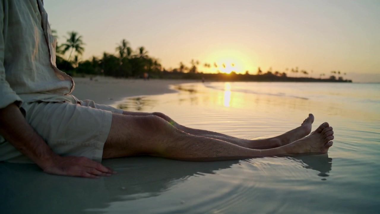 Ola entrante haciendo que un hombre se mueva y flexione los dedos de los pies en una playa de arena, con el resplandor del sol, camiseta de lino y espacio para texto.