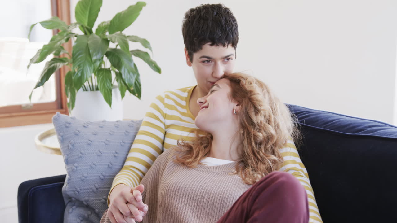 Happy caucasian lesbian couple sitting,,embracing and holding hands on sofa in sunny house