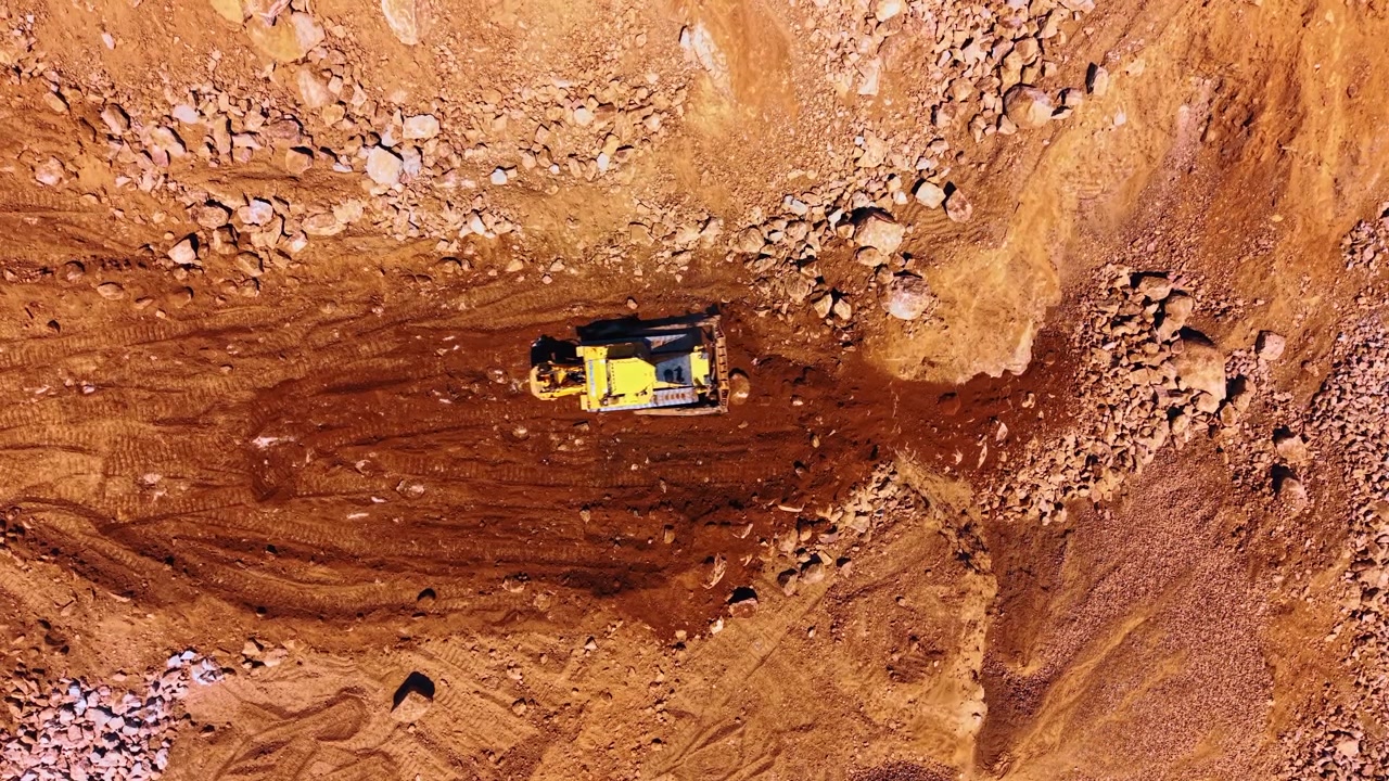 Top down view of bulldozer in stone quarry. Yellow crawler bulldozer moving dark soil and rocks on brown ground surface.