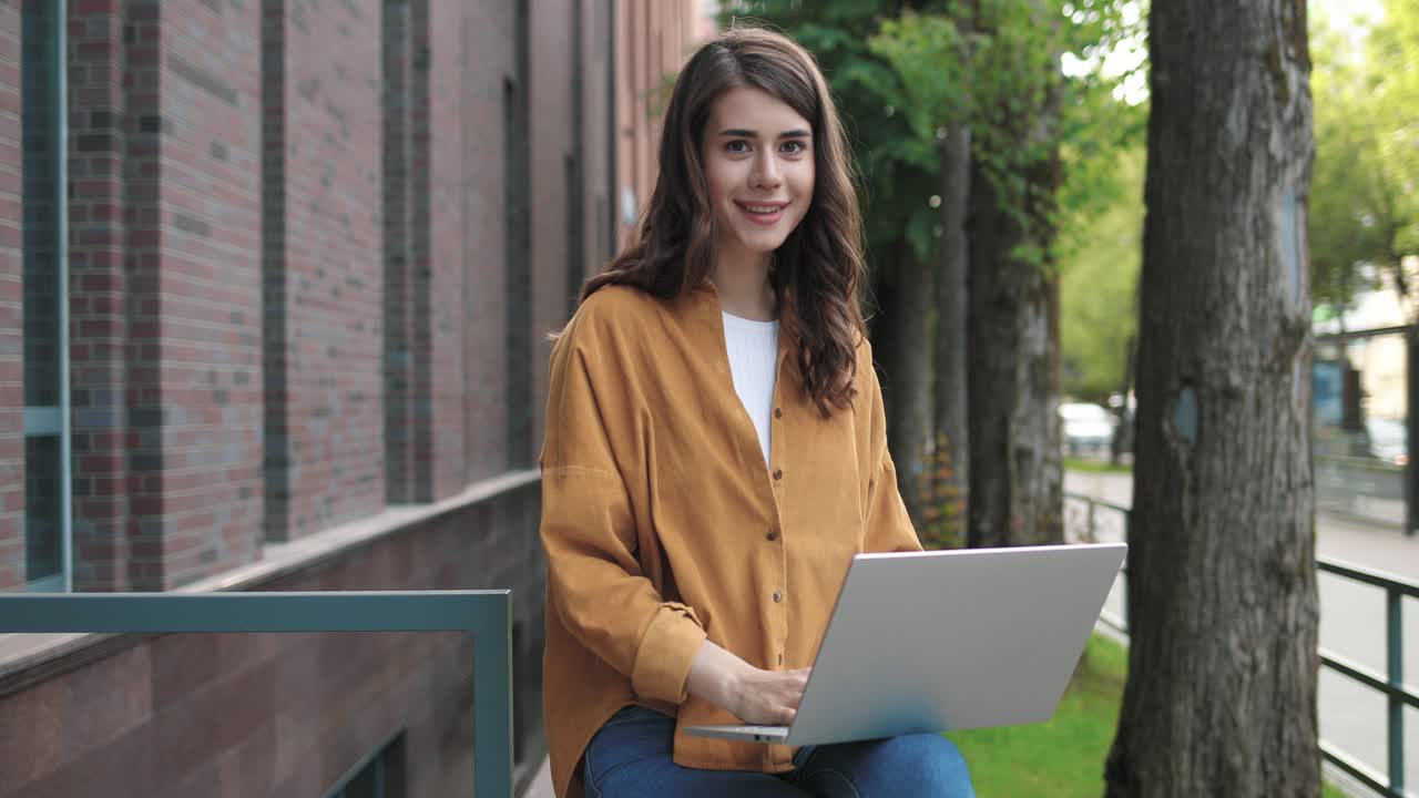 cámara haciendo zoom en una estudiante caucásica sentada en la calle y sonriendo a la cámara mientras usa una laptop cerca de la universidad