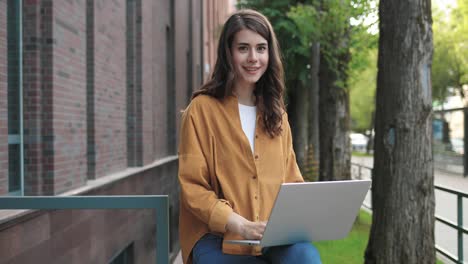 camera zooming on caucasian female student sitting in the street and smiling to the camera while using a laptop near the college