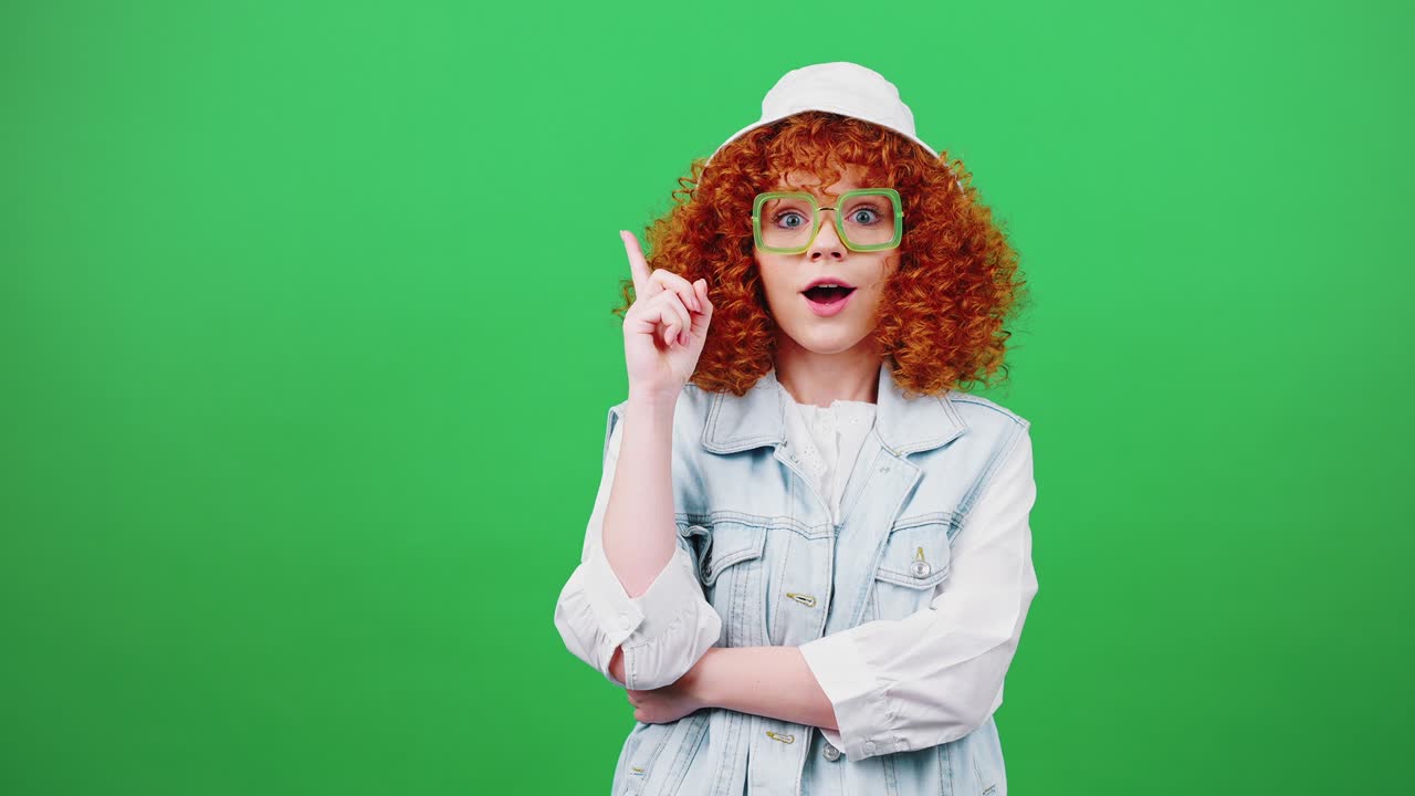 joven roja bonita mujer pensando, teniendo idea y señalando con el dedo hacia arriba, riendo sobre el fondo clave de croma verde