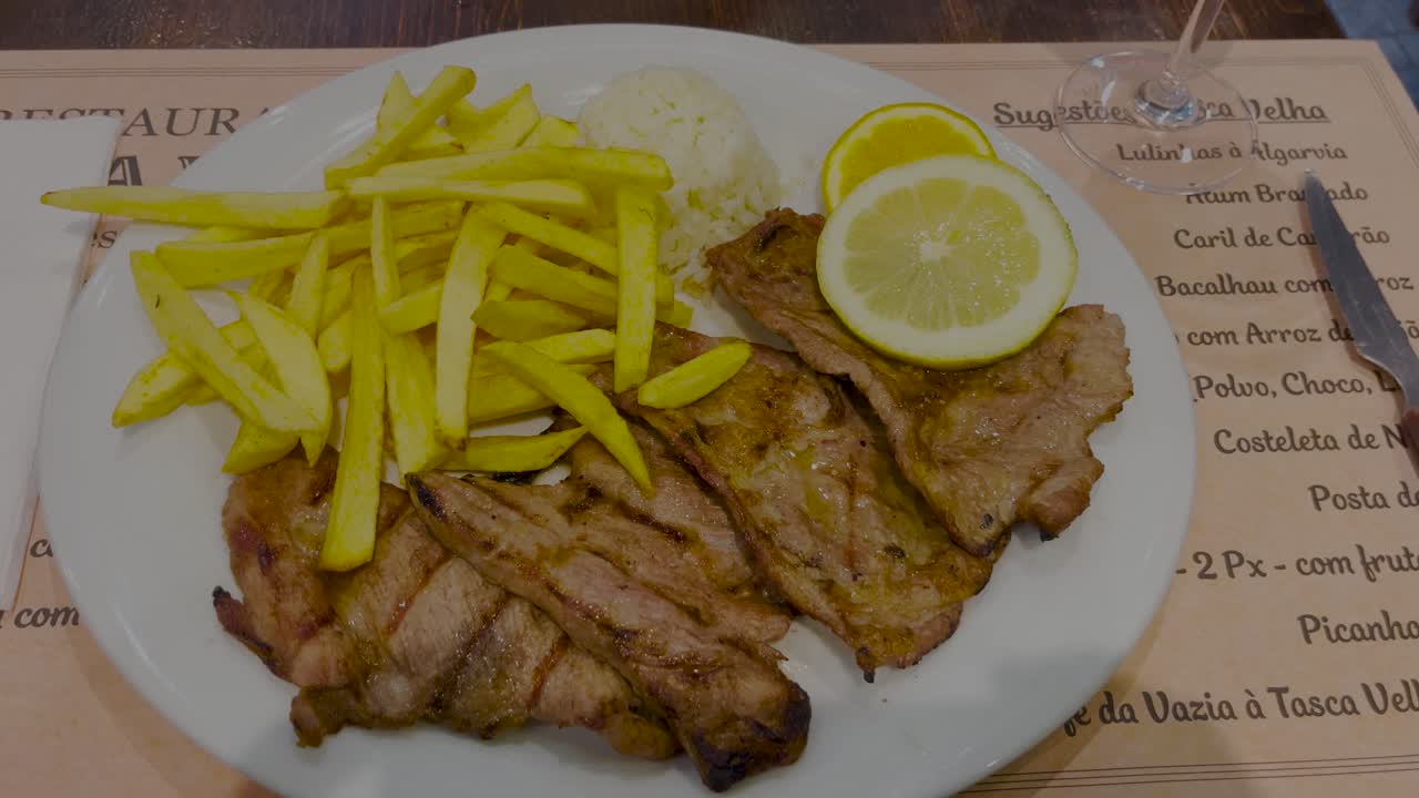 A plate of delicious grilled pork with fries and rice, garnished with fresh lemon slices, offering a satisfying and hearty meal that blends savory flavors and comfort food.