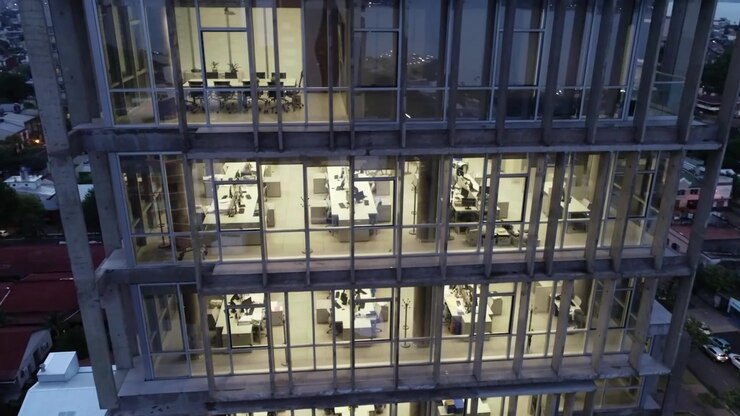 Escritorios de oficina brillantemente iluminados visibles a través de grandes ventanales de cristal de un rascacielos de hormigón