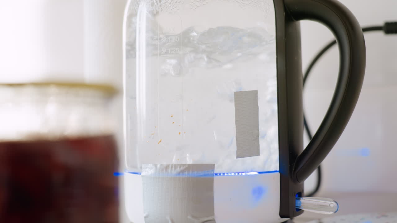 Close up of electric kettle rapidly boiling water with visible steam and bubbles plastic handle in focus, blue light glowing at base, background slightly blurred