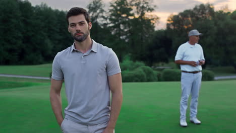 confident man standing golf course outside. two golfers relax at sunset field.