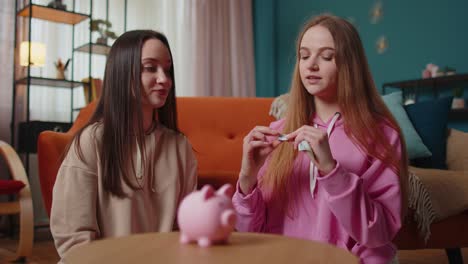 Girls-friends-siblings-sitting-on-floor-and-take-turns-dropping-dollar-banknote-into-piggy-bank