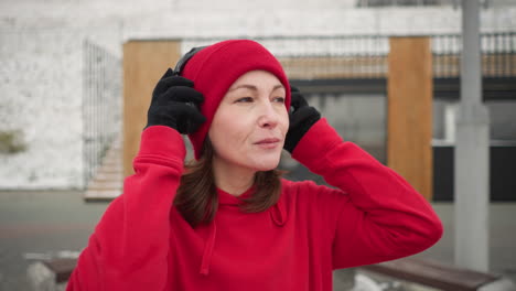 middle aged woman wearing red beanie and hoodie smiles while putting on headphones outdoors in winter, gazing into the distance, background features modern architecture and snow-covered urban setting