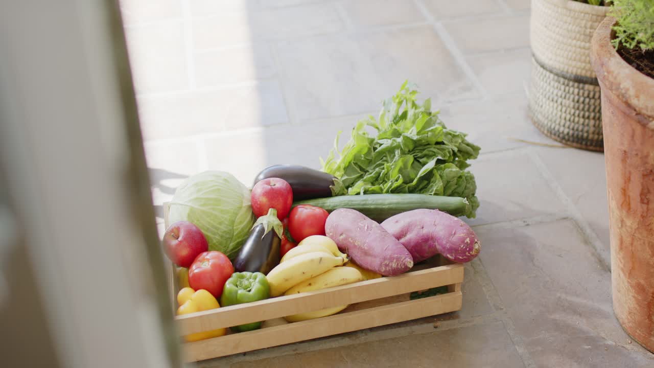 Cámara deslizándose hacia adentro, revelando una caja de alimentos con plátanos y tubérculos, la luz del sol resaltando los productos