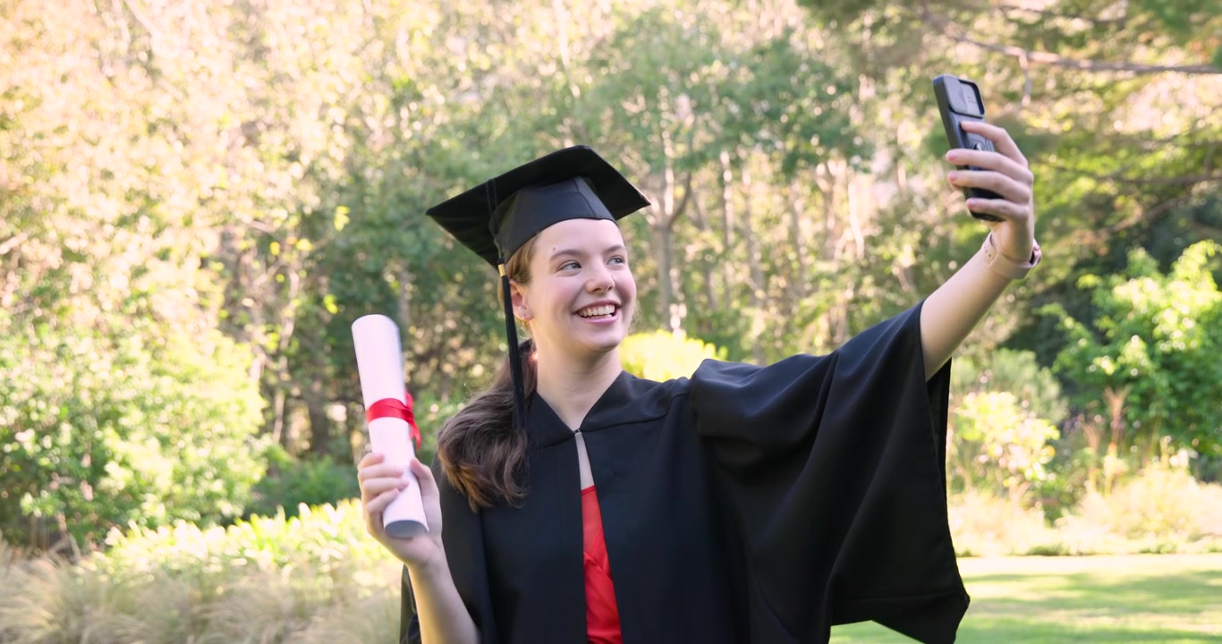 Laureata con tocco che festeggia la laurea al parco, scattandosi selfie con il telefono e sventolando il diploma.