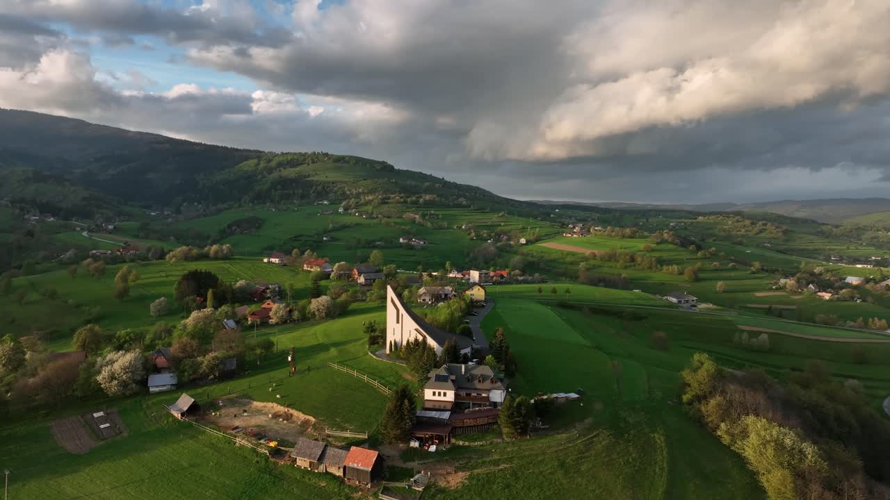 un avión no tripulado orbita y revela una iglesia moderna que corona una colina iluminada por el sol en una tranquila noche de primavera, enmarcada por un espectacular dosel de nubes, un cielo azul cristalino y la cálida luz del sol