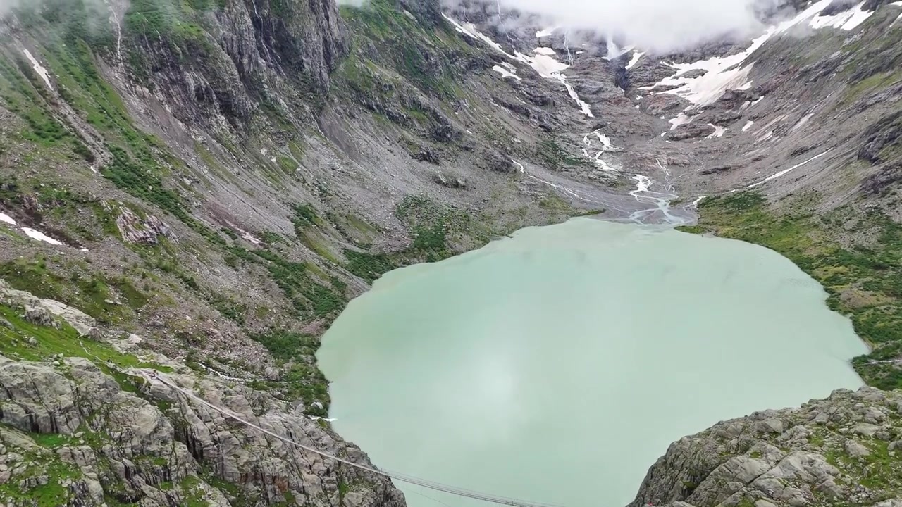 Ripresa aerea in tilt-up verso il Triftbrücke in Svizzera, che enfatizza la bellezza alpina e la struttura del ponte sospeso alla base del lago.