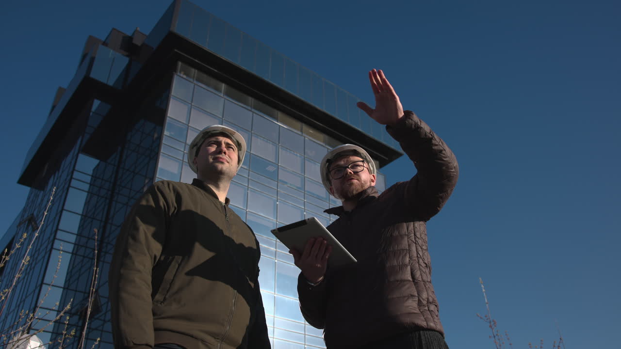 Due professionisti dell'edilizia in cantiere indicano la facciata in vetro, un ingegnere tiene un tablet