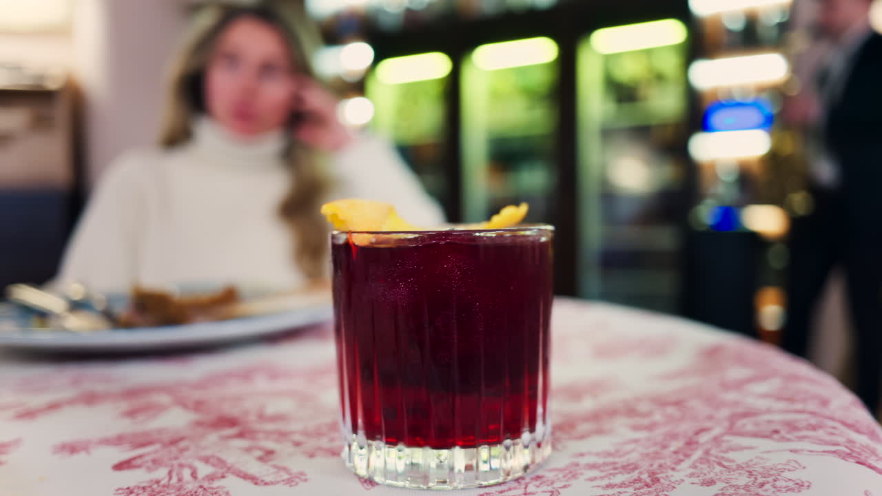 Primo piano di un cocktail negroni su una tovaglia rossa e bianca con una donna seduta in un ristorante sullo sfondo