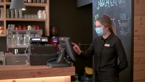 a waitress with face mask checks the orders on a screen next to the bar counter