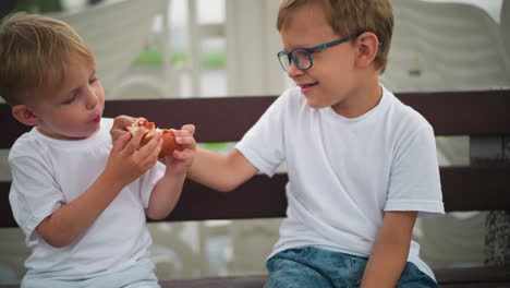 two young siblings sit on a bench outdoors, with the older child reaching out to take some snacks from the younger sibling