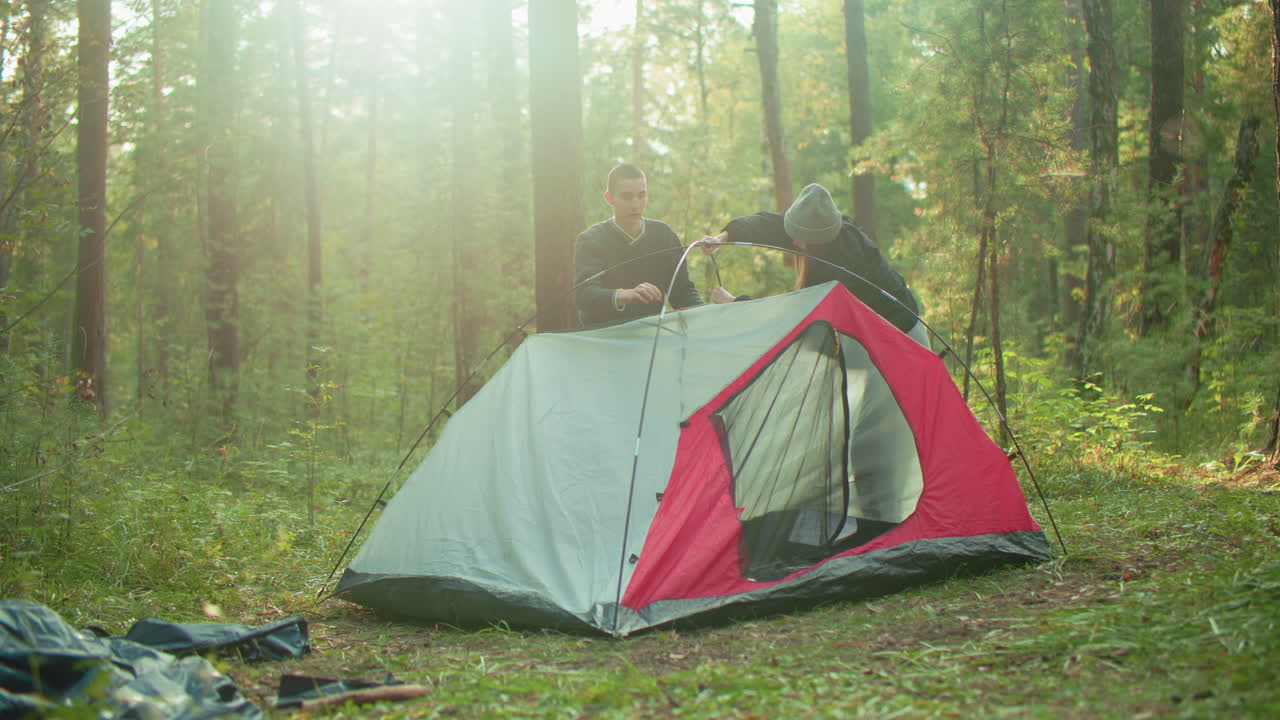 young couple stand on opposite sides of pitched tent in forest as sunlight glows through trees, surrounded by scattered camping items while working together to complete tent setup