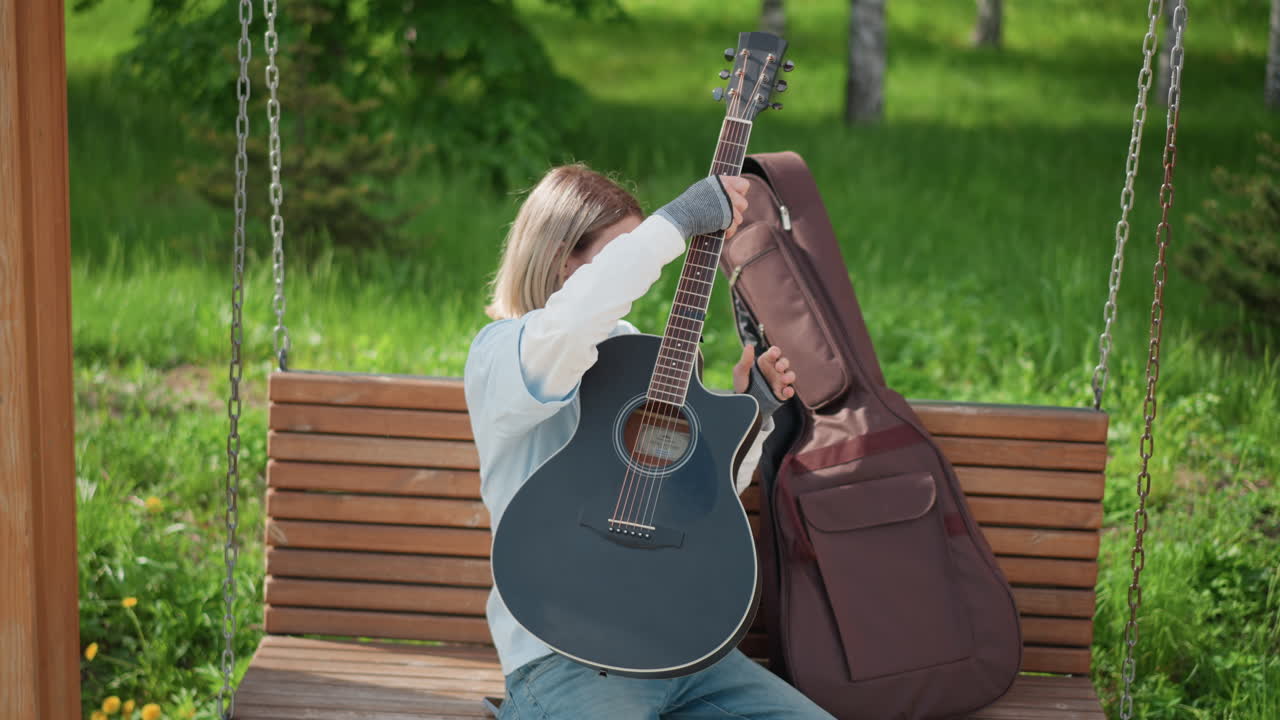 Joven guitarrista afinando las cuerdas de su guitarra en un banco del parque, probando acordes y ajustando las clavijas, sacando la guitarra de su funda, rasgueando una melodía suave, sesión de práctica al aire libre con luz solar y hierba